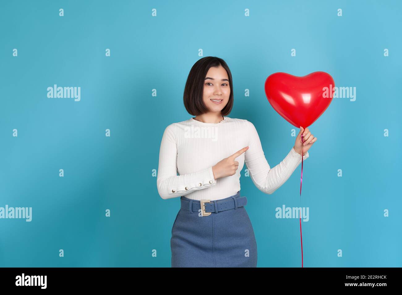 Una giovane donna asiatica gioiosa e impressionata tiene un pallone volante a forma di cuore e lo punta con il dito indice , isolato su uno sfondo blu Foto Stock