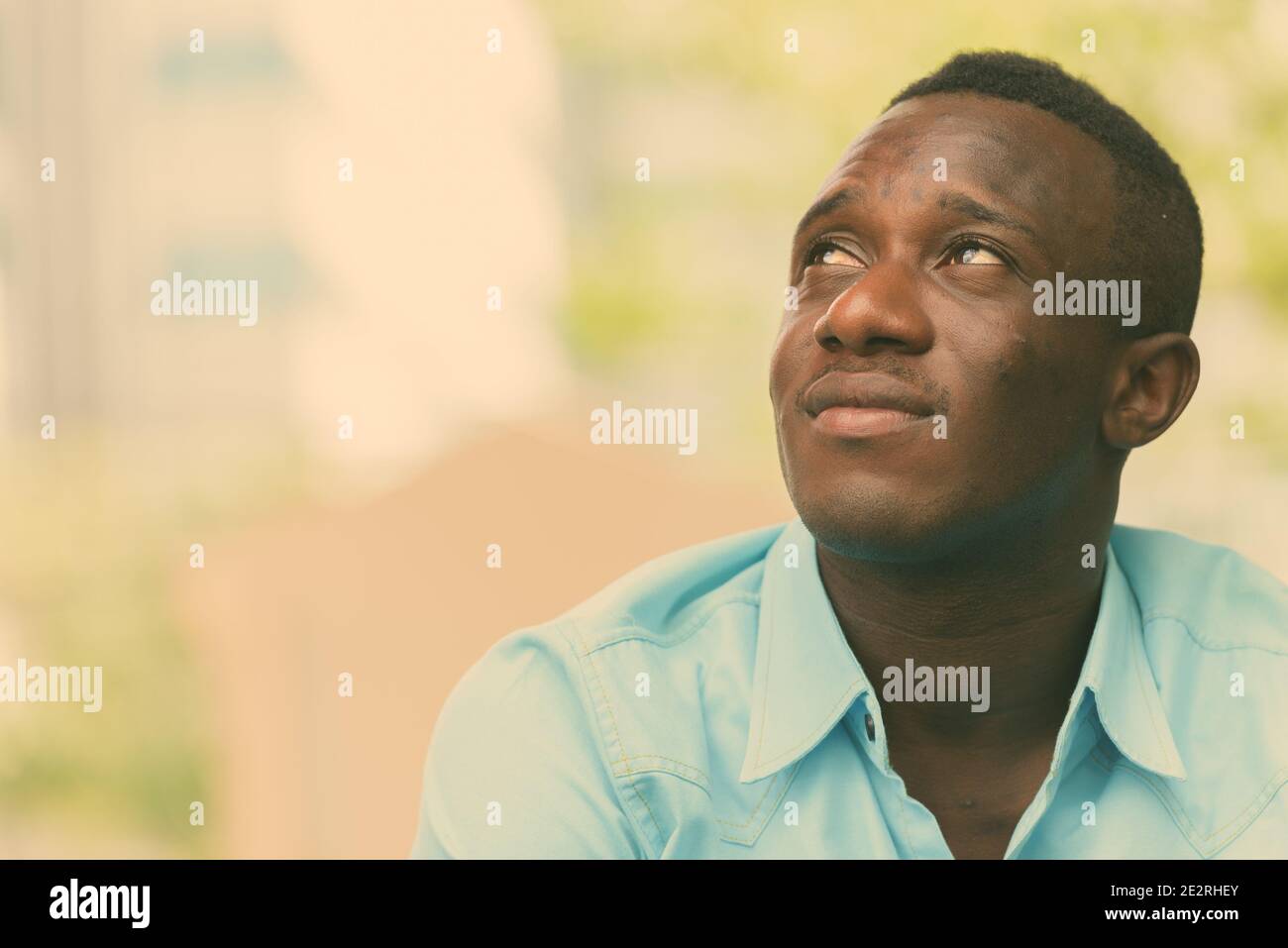 Giovane Africano nero uomo seduto mentre il pensiero e la ricerca al di fuori del coffee shop contro la vista della natura e della città Foto Stock