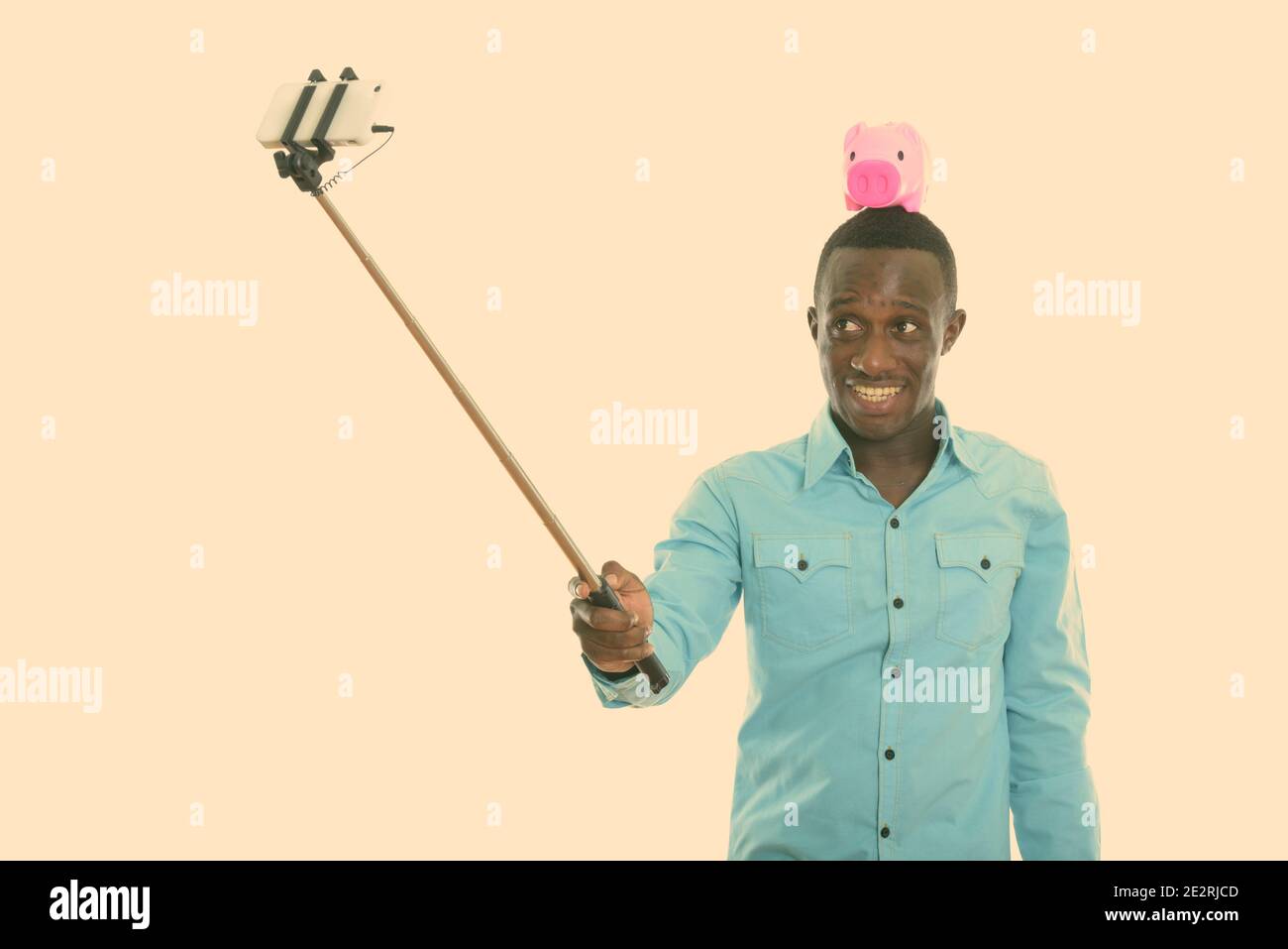 Studio shot di giovani felici Africano nero uomo sorridente con il salvadanaio sulla testa tenendo selfie foto con il telefono cellulare su selfie stick Foto Stock