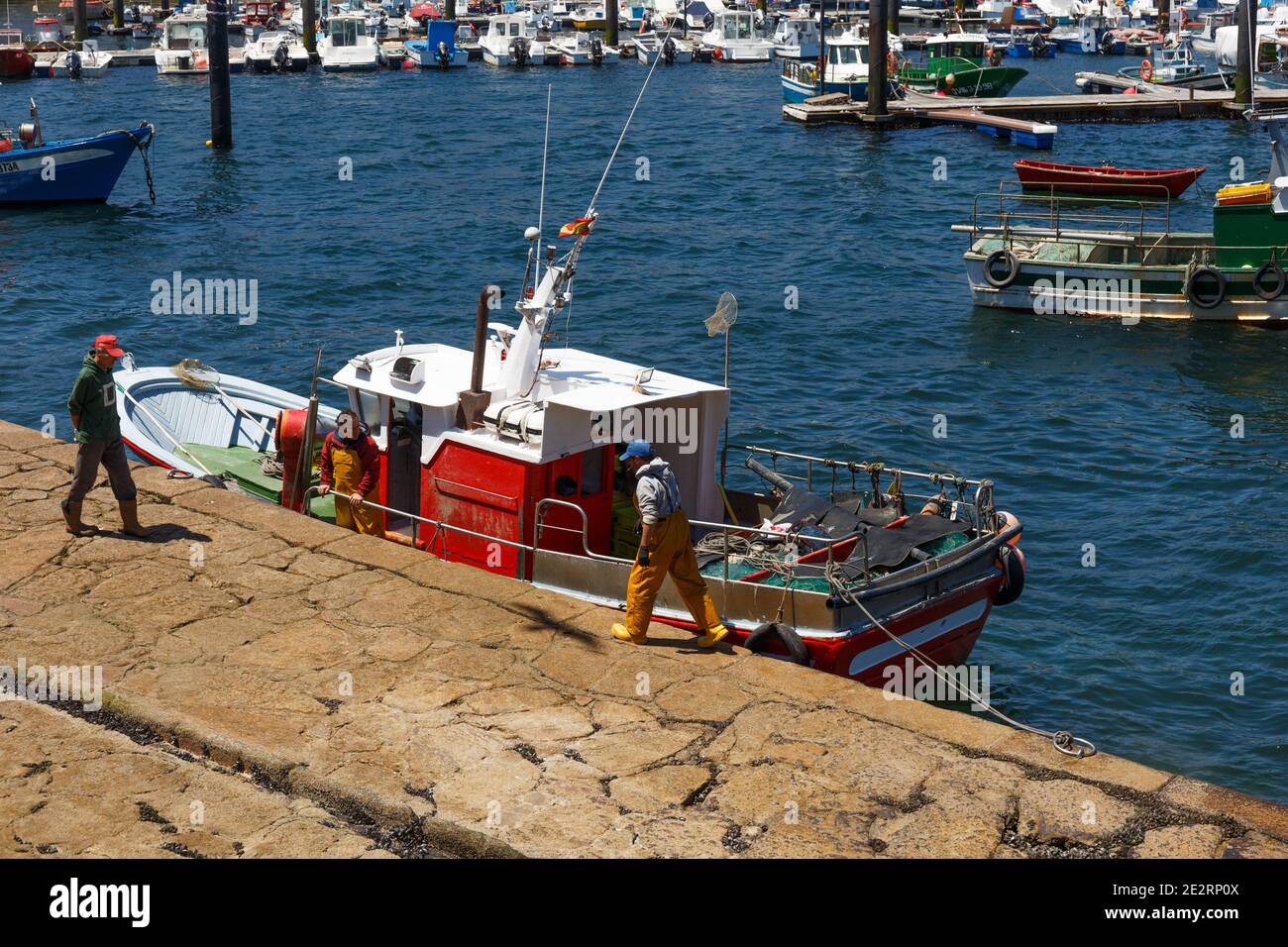 Barca da pesca da vicino, due pescatori e una persona a piedi, con vista sulla città sullo sfondo in una giornata di sole, Finisterre, Galizia, Spagna Foto Stock