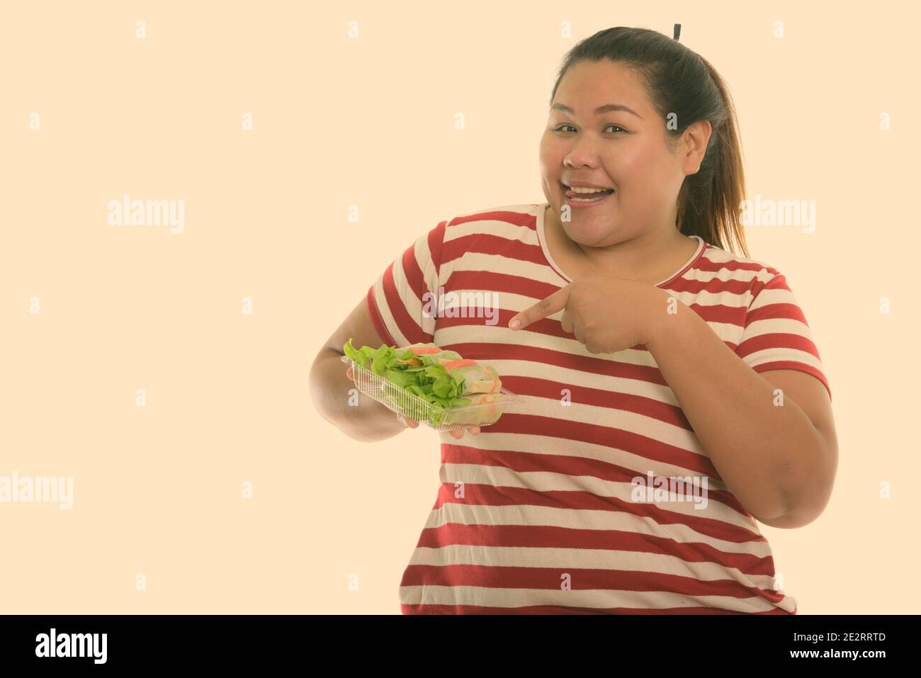 Studio shot di giovani felici fat donna asiatica sorridere mentre holding e puntando alla verdura fresca avvolgere mentre con la lingua fuori Foto Stock