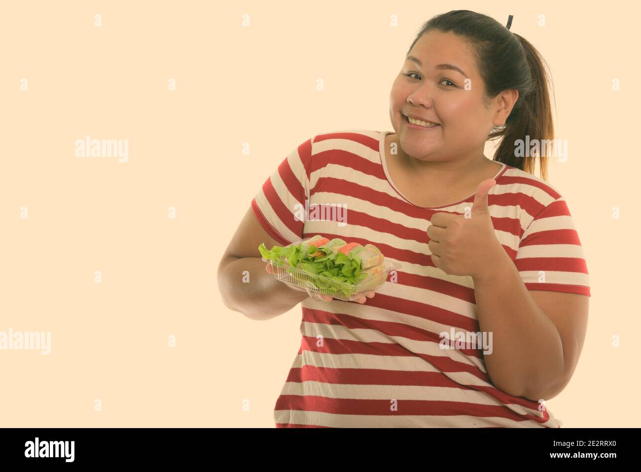 Studio shot di giovani felici fat donna asiatica sorridere mentre dando pollice in alto e tenendo premuto verdura fresca wrap Foto Stock