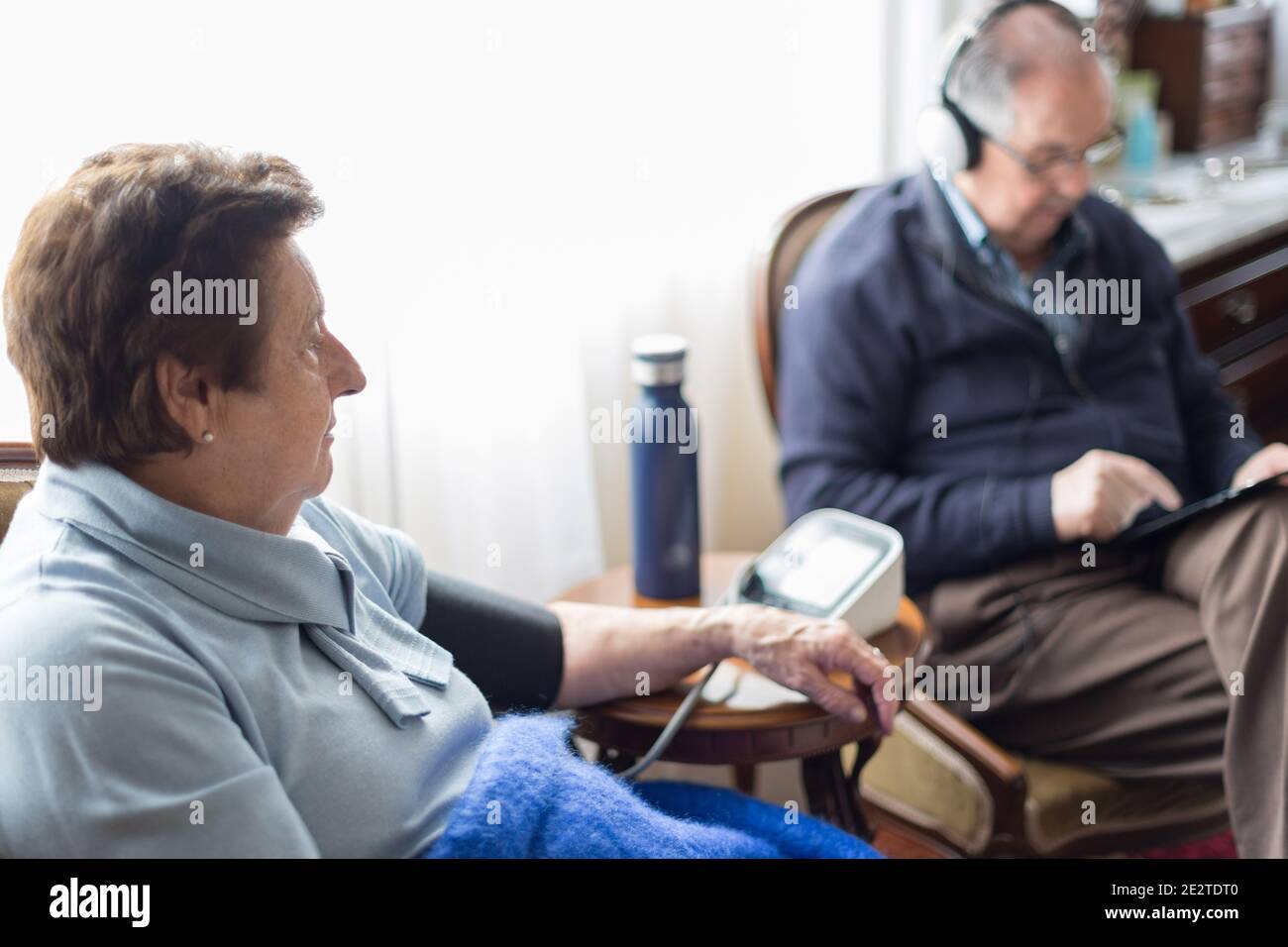 Una coppia di anziani caucasici a casa utilizzando dispositivi digitali e controllando la pressione sanguigna vestita di blu. Foto Stock