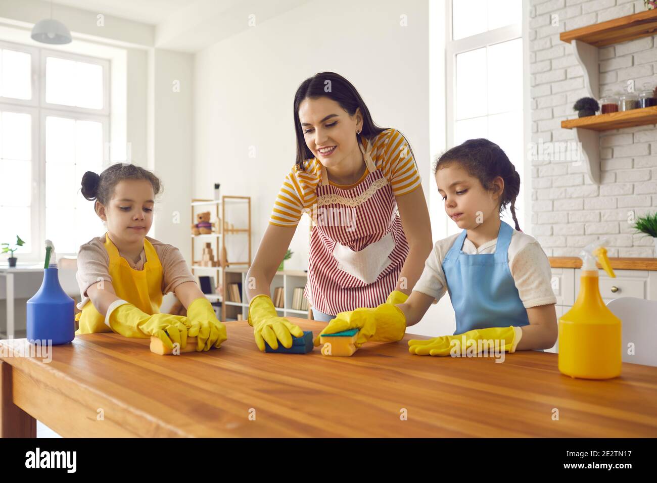 Felici piccole figlie che aiutano la loro mamma a lavare la tavola con spugne e detersivo Foto Stock