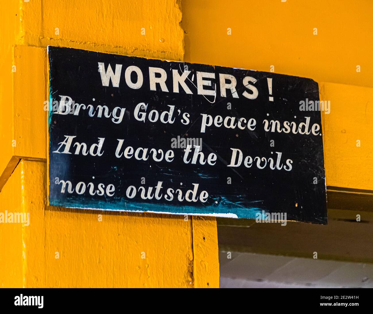 “Lavoratori! Portate la pace di Dio all’interno. E lasciate il rumore del diavolo fuori.” recita un cartello all’interno della fabbrica di noce moscata a Gouyave, Saint John, Grenada. L’appello ai lavoratori è appeso a un palo di legno dipinto di giallo all’interno dell’edificio. Il paese è il secondo più grande produttore mondiale di noce moscata dopo l’Indonesia. Foto Stock