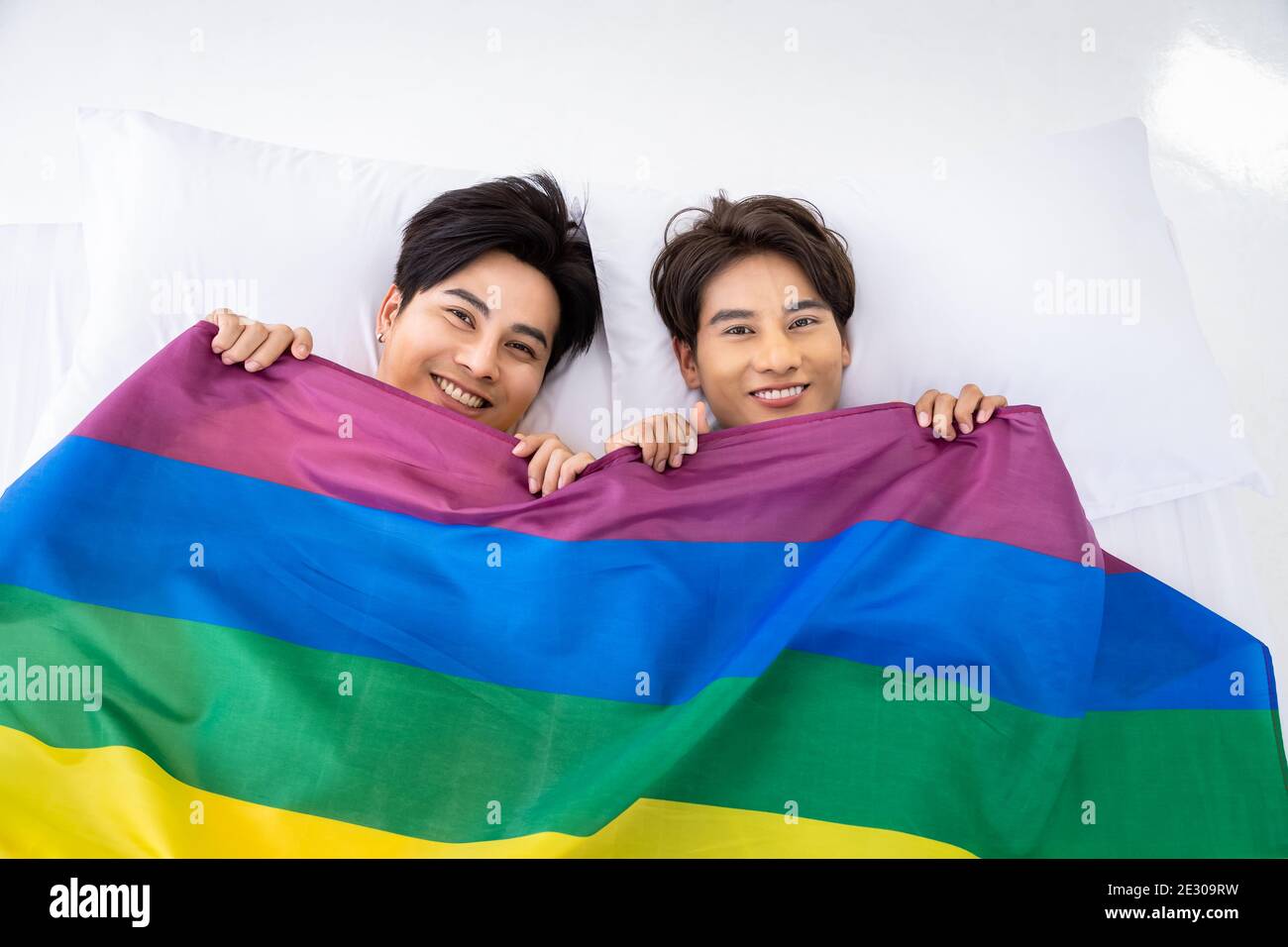 Felice coppia omosessuale asiatica giace su letto bianco con una copertura di bandiera di orgoglio arcobaleno, guardando la macchina fotografica. Concetto LGBT Foto Stock
