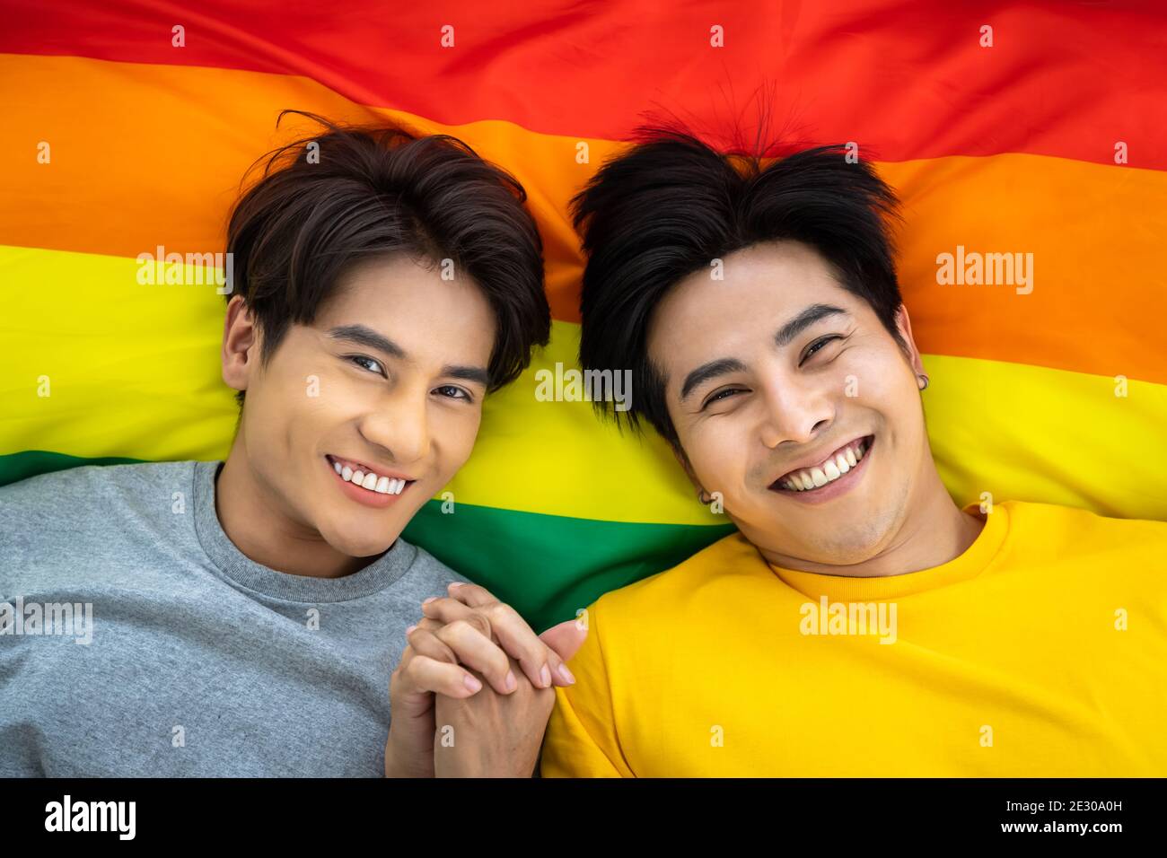 Felice coppia omosessuale asiatica che giace sulla bandiera dell'orgoglio iridato, tenendo la mano. Concetto LGBT. Guardando la fotocamera. Foto Stock