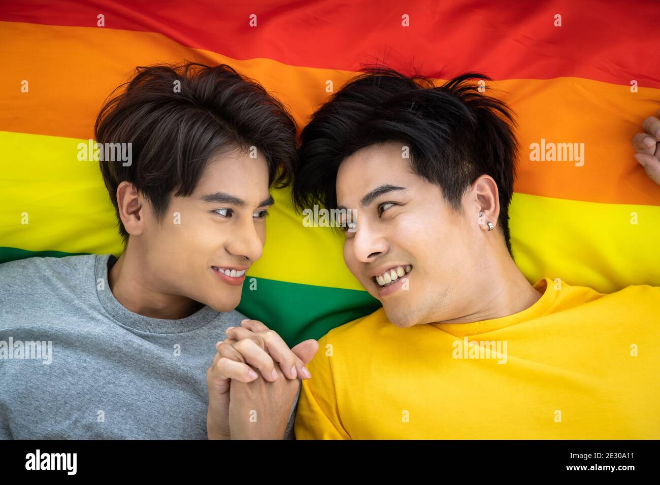 Felice coppia omosessuale asiatica che giace sulla bandiera dell'orgoglio iridato, tenendo la mano. Concetto LGBT. Foto Stock