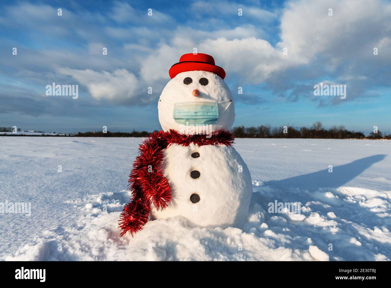 Simpatico pupazzo di neve in elegante cappello rosso con maschera medica su campo innevato. Concetto di allontanamento sociale e di viaggio annullato Foto Stock