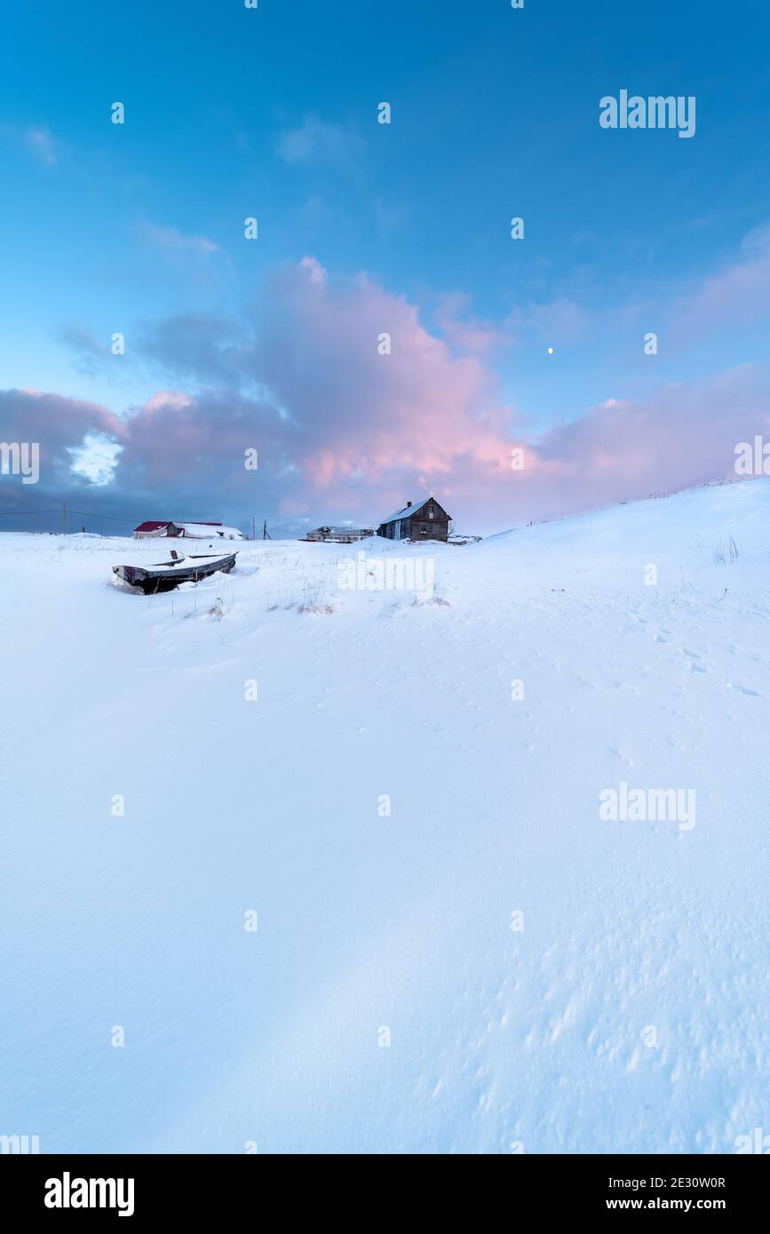 Barca di legno e casa di legno sul campo innevato in un paesaggio artico al tramonto con la luna nel cielo Foto Stock