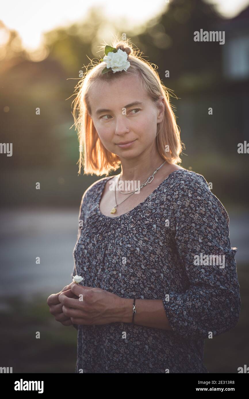 Una donna dai capelli biondi indossa un fiore bianco nei capelli, retroilluminato dal sole che tramonta durante l'ora d'oro Foto Stock