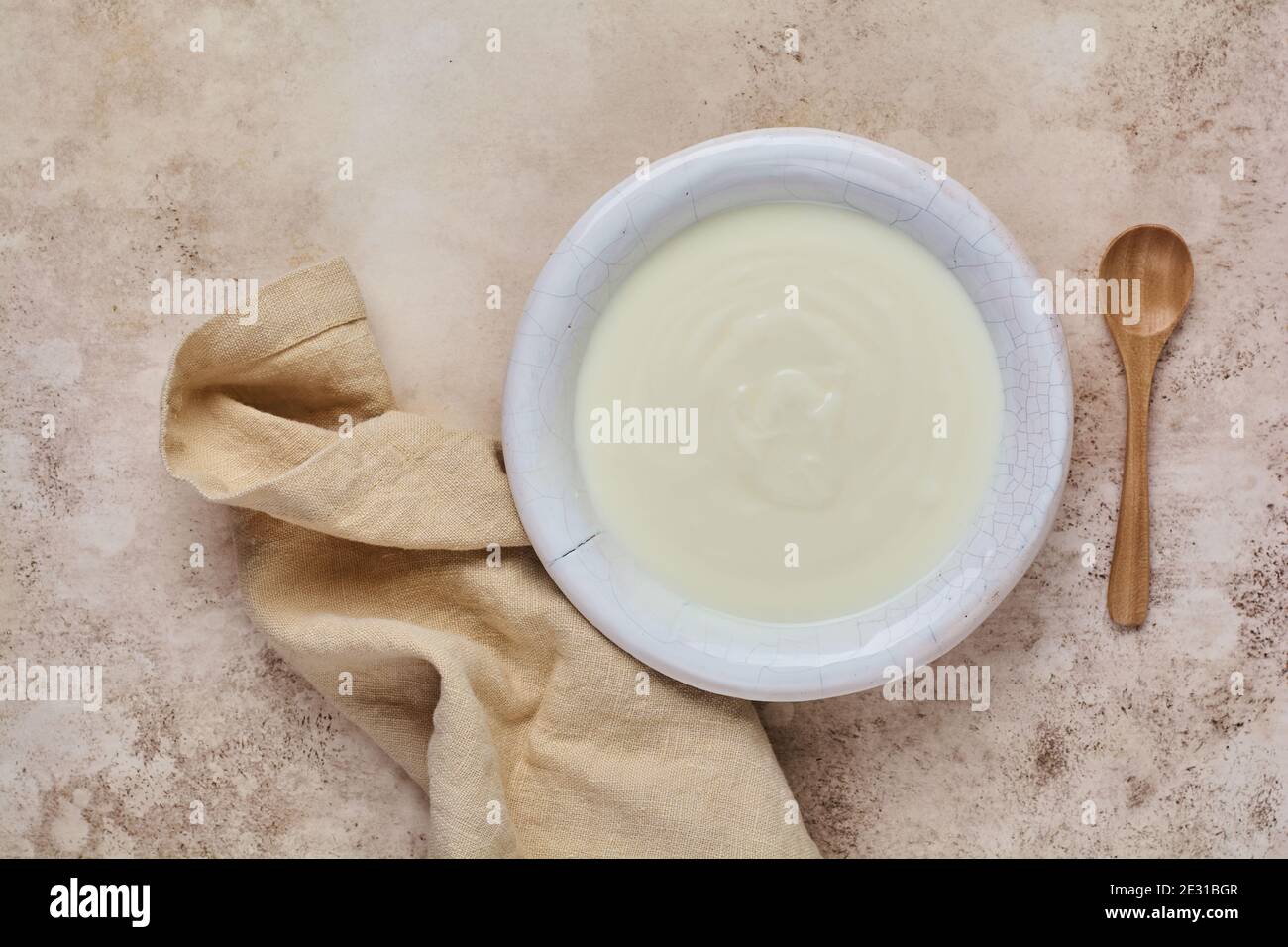 Yogurt greco in ciotola bianca, tovagliolo di lino e cucchiaio di legno su vecchio tavolo beige. Foto Stock