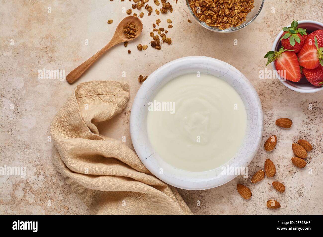 Yogurt greco in ciotola bianca con ingredienti per preparare la colazione a base di muesli e fragole fresche su vecchio tavolo beige. Foto Stock