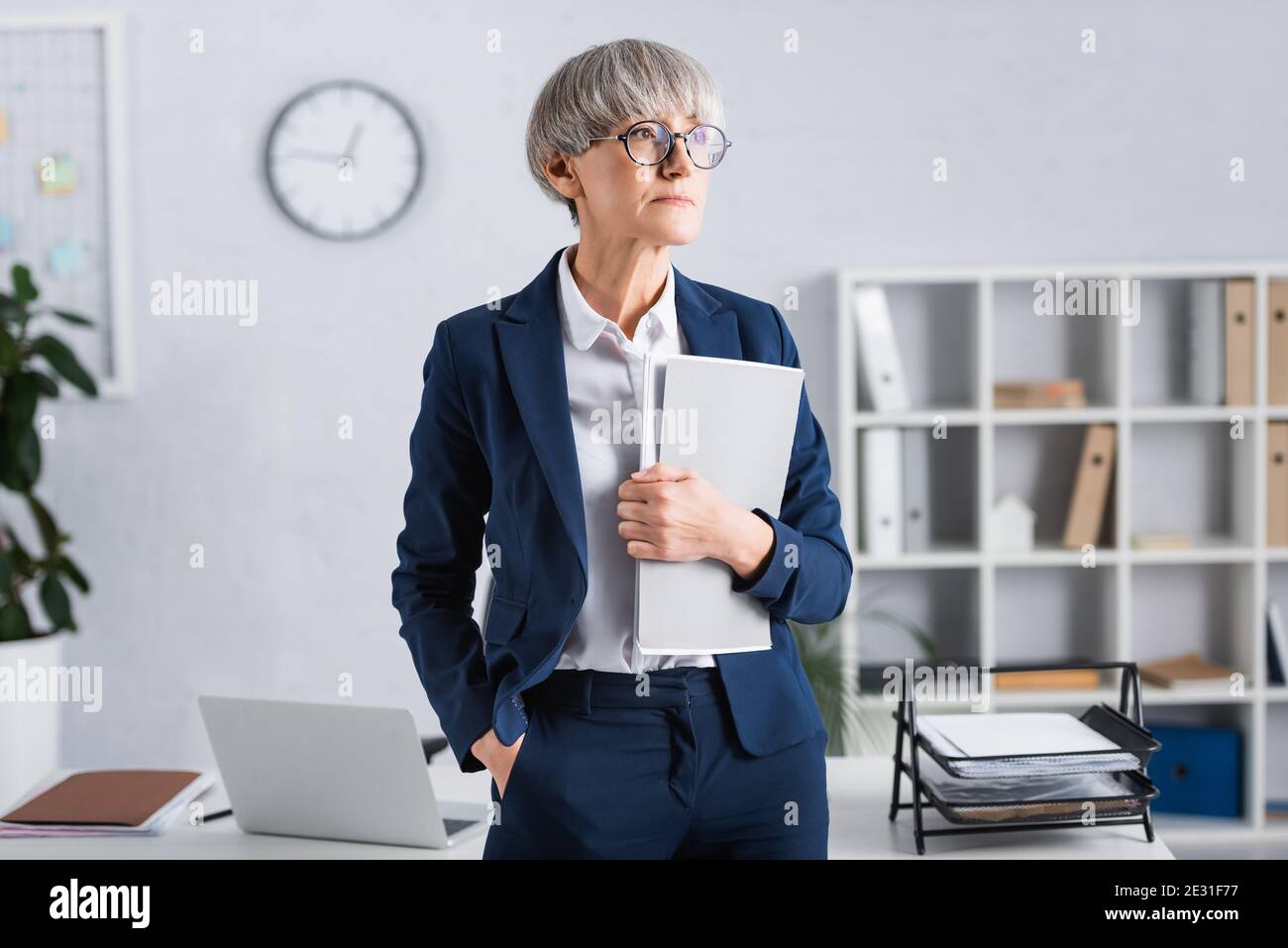 leader del team di media età in occhiali e tuta in piedi porta in tasca e tieni la piegatrice in ufficio Foto Stock