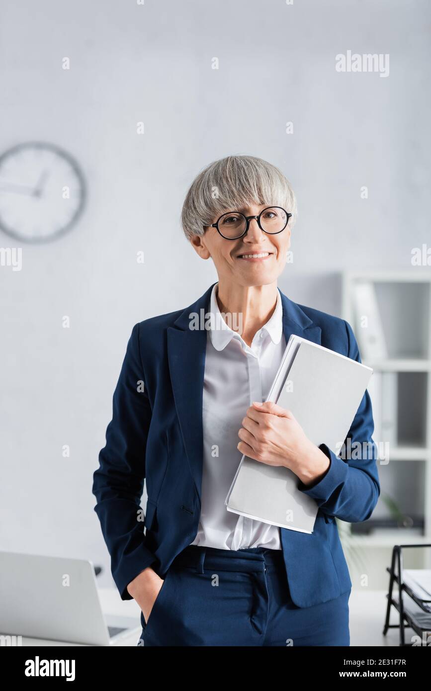 felice team leader di mezza età in occhiali e tuta in piedi con tasca a mano e piegatrice in ufficio Foto Stock