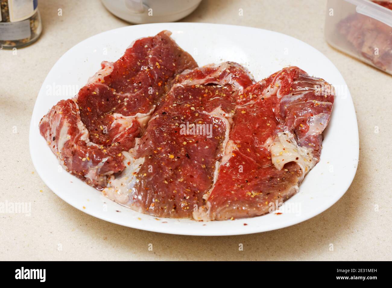 Pezzi di carne cruda cosparsi di spezie su piatto bianco prima di friggere Foto Stock
