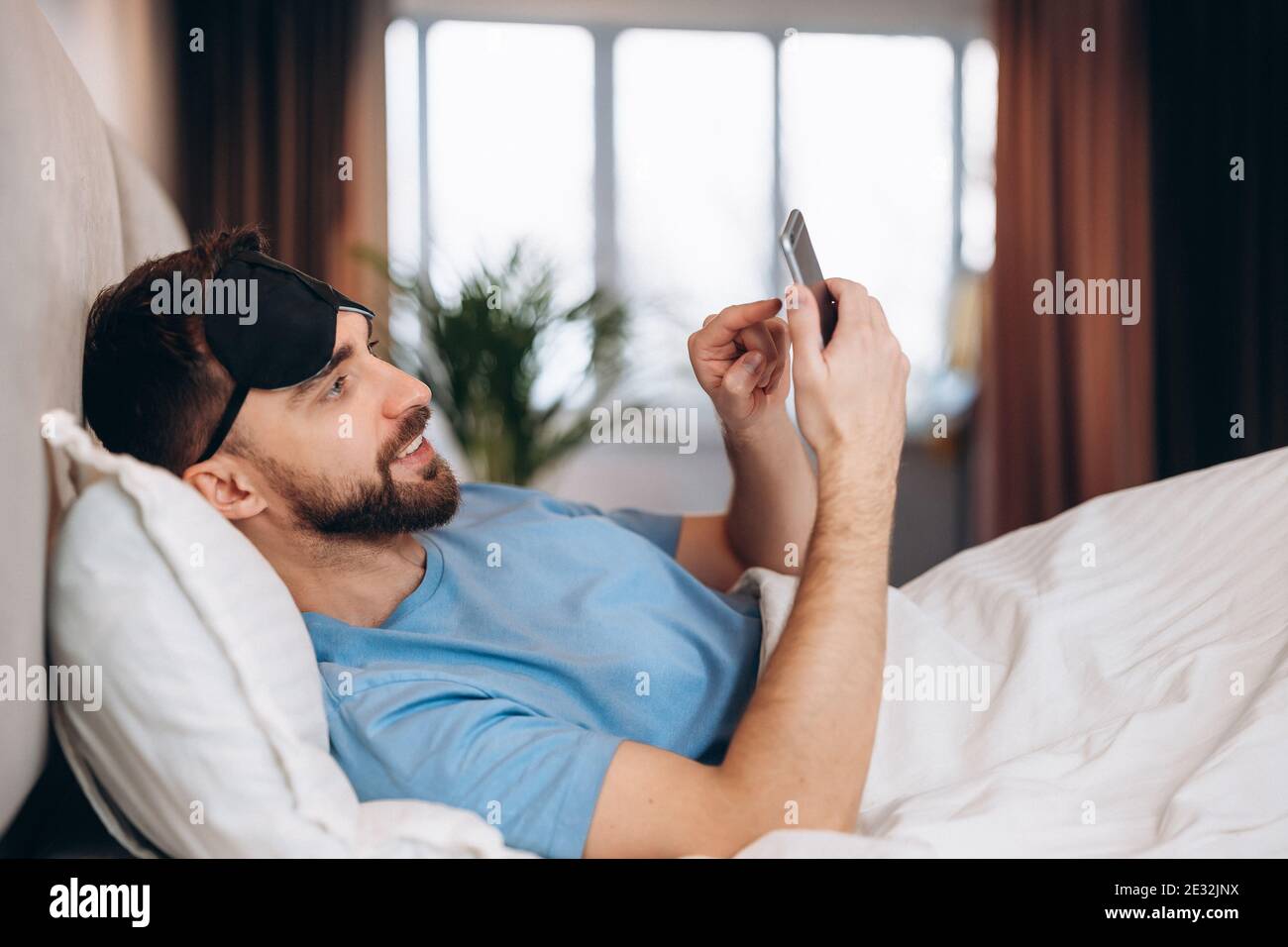 Giovane attraente uomo sdraiato a letto controllando i suoi messaggi sul suo telefono al mattino pigro giorno fuori. Foto Stock