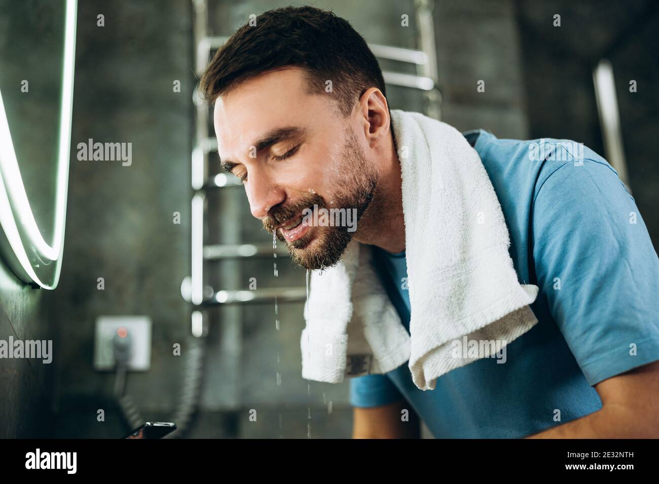 Giovane uomo con barba che si lava il viso. Persona pulizia viso con asciugamano sul collo sapone in bagno lavabo a casa Foto Stock