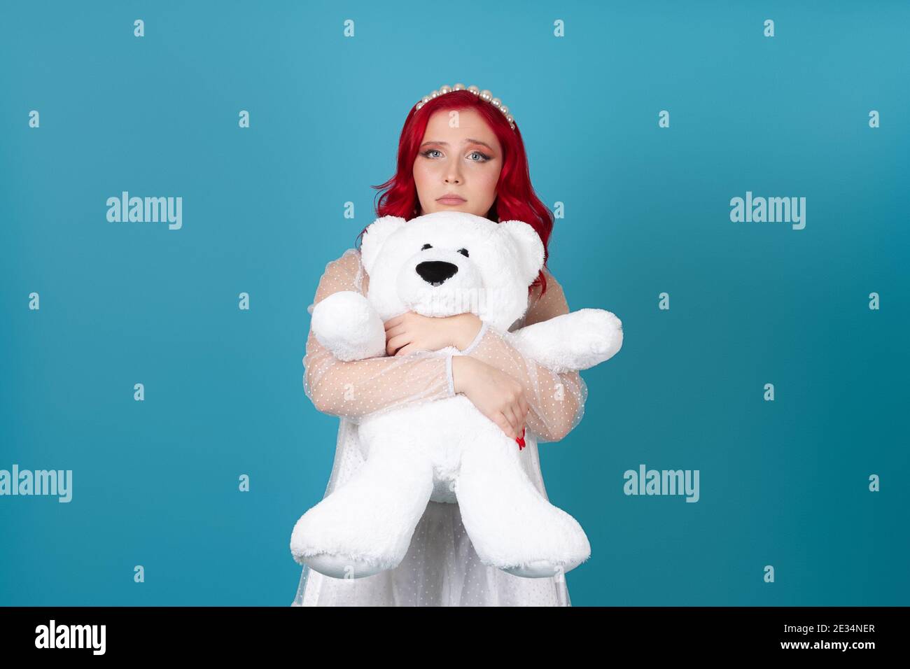 triste, ferito, deluso, una donna in un vestito bianco con capelli rossi abbraccia delicatamente un orsacchiotto bianco isolato su uno sfondo blu Foto Stock