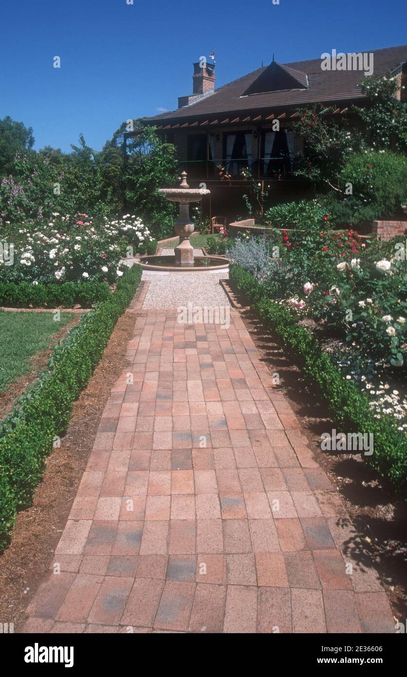 Percorso del giardino di mattoni bordato con basse piante di copertura e che conduce ad una grande fontana. Il giardino presenta cespugli di rose e piante assortite. Foto Stock