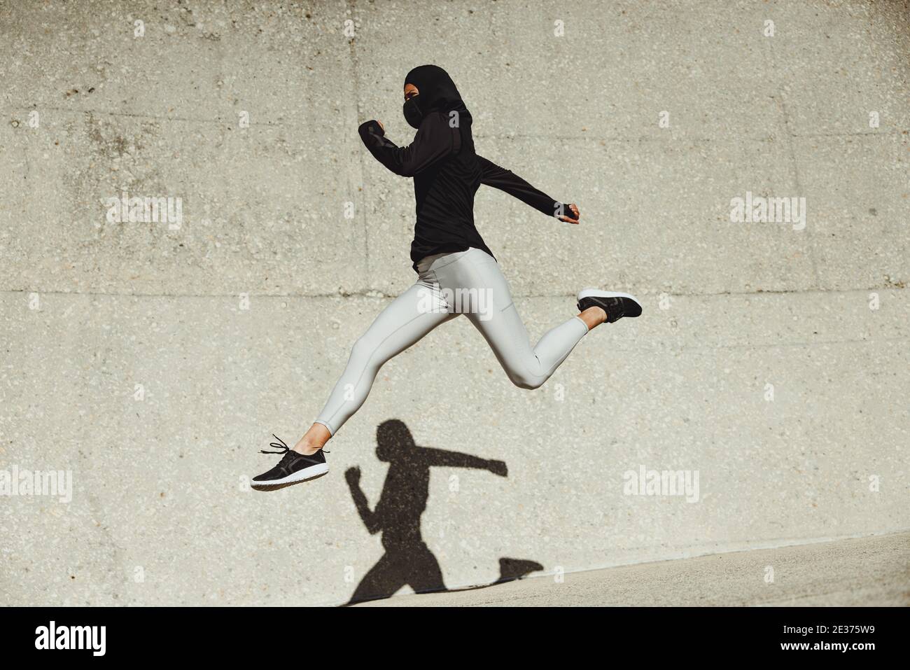 Donna che indossa l'abbigliamento da training hijab e maschera protettiva per il viso che si esercita all'aperto. Donna che corre e salta in abbigliamento sportivo. Foto Stock