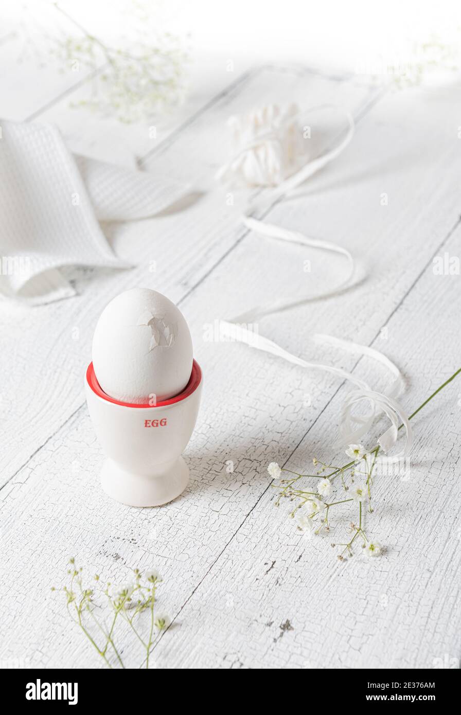 Vita morta creativa stagionale con uova di pollo cracked sul supporto dell'uovo, fiori bianchi freschi, tovagliolo tessile e nastro bianco su t di legno bianco Foto Stock