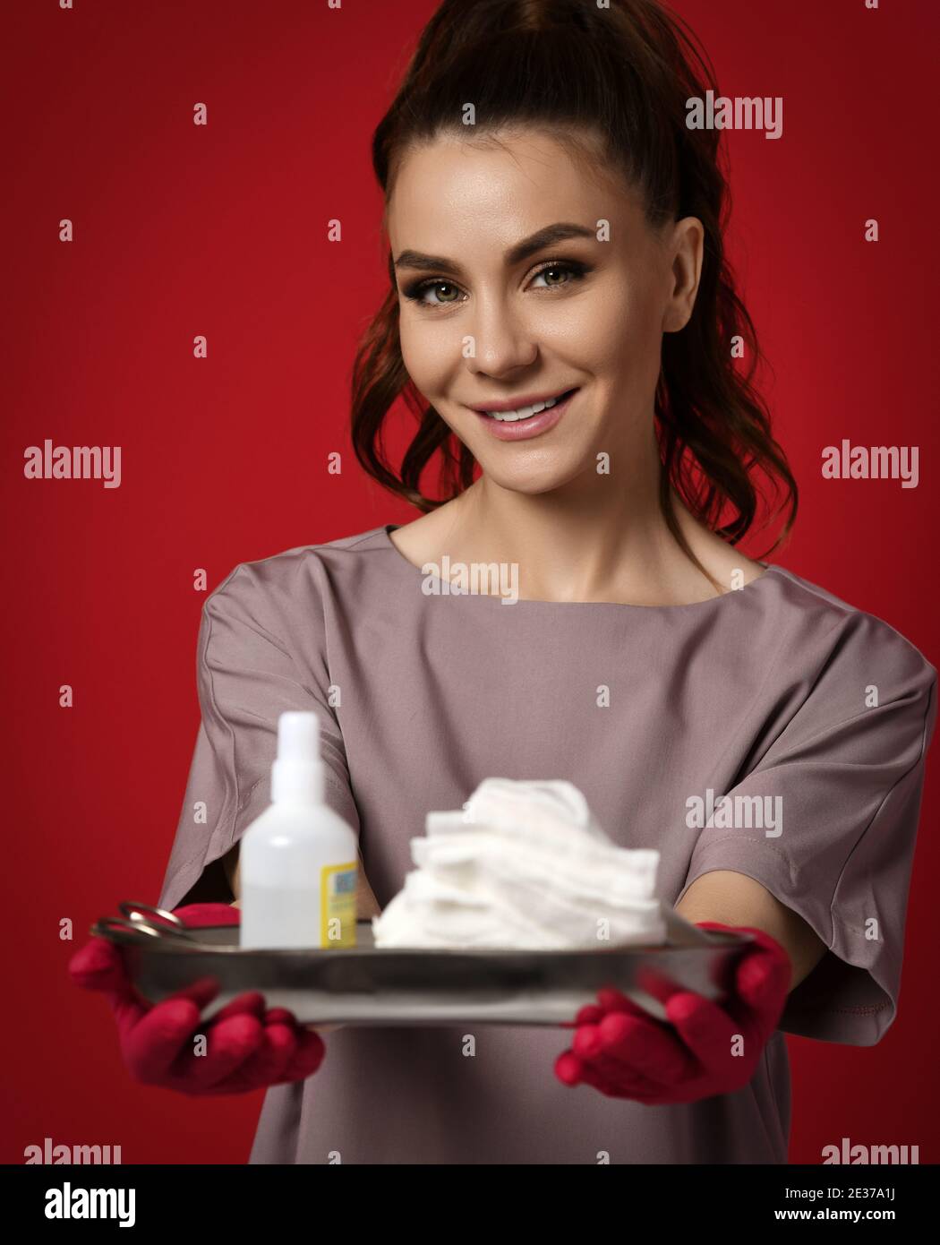 Sorridente bruna bella donna infermiere medico in rosso guanti di lattice contiene strumenti medici, antisettici, bendaggio, garza su vassoio Foto Stock
