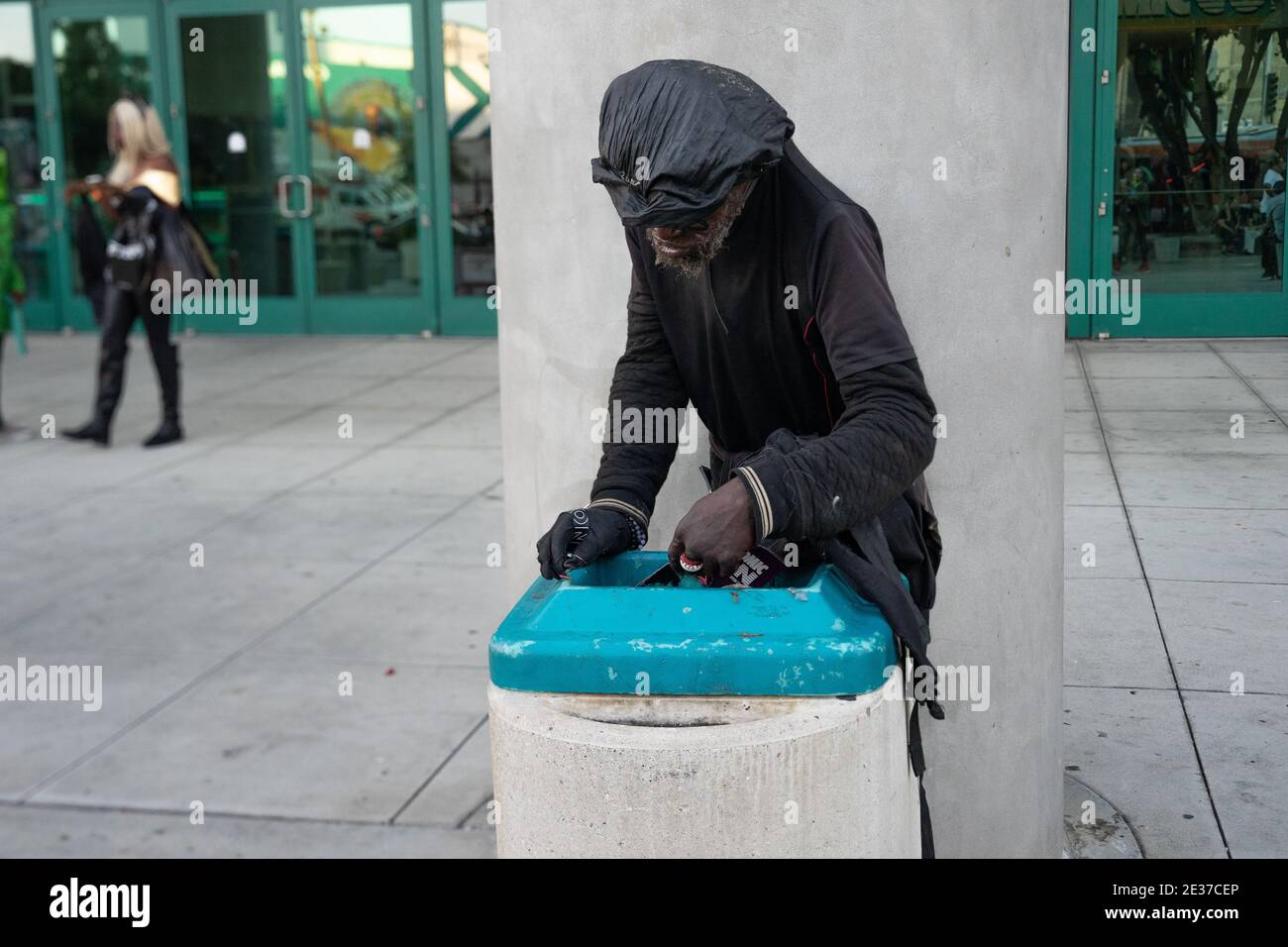 Un uomo nero vestito di outerwear nero sporco raccoglie un opuscolo da un contenitore di rifiuti a Los Angeles, CA, Stati Uniti Foto Stock