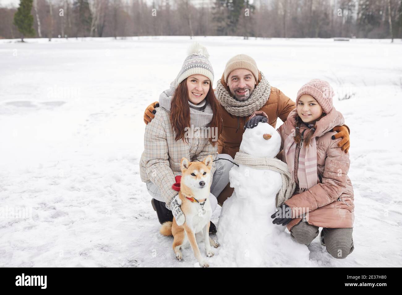 Ritratto a lunghezza intera di felice famiglia che costruisce pupazzo di neve insieme all'aperto e guardando la macchina fotografica mentre godendo la vacanza invernale, copy space Foto Stock