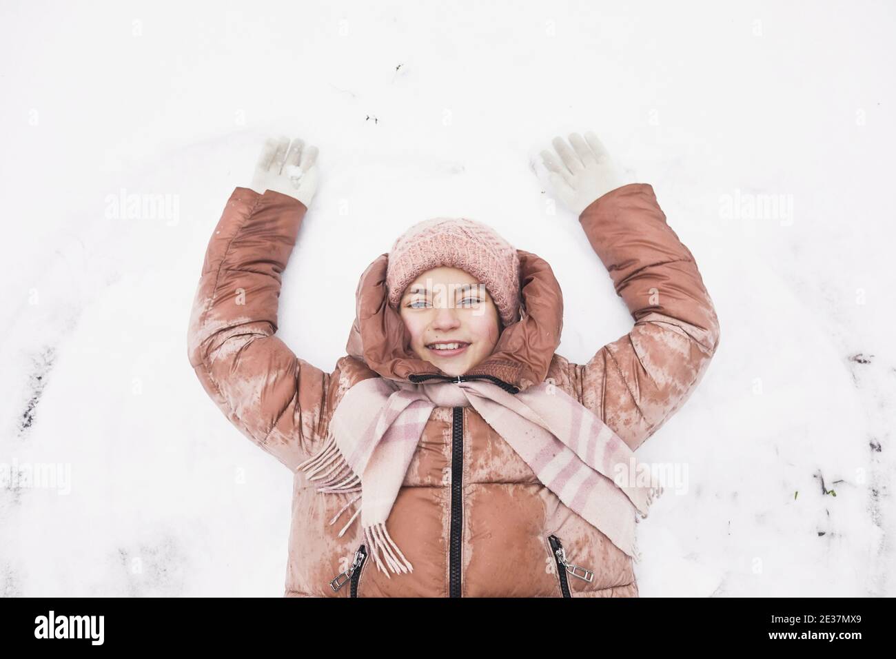 Vista dall'alto verso il basso della ragazza carina sdraiata nella neve e facendo angeli di neve mentre si gode l'inverno all'aperto e sorridendo alla macchina fotografica, copia spazio Foto Stock