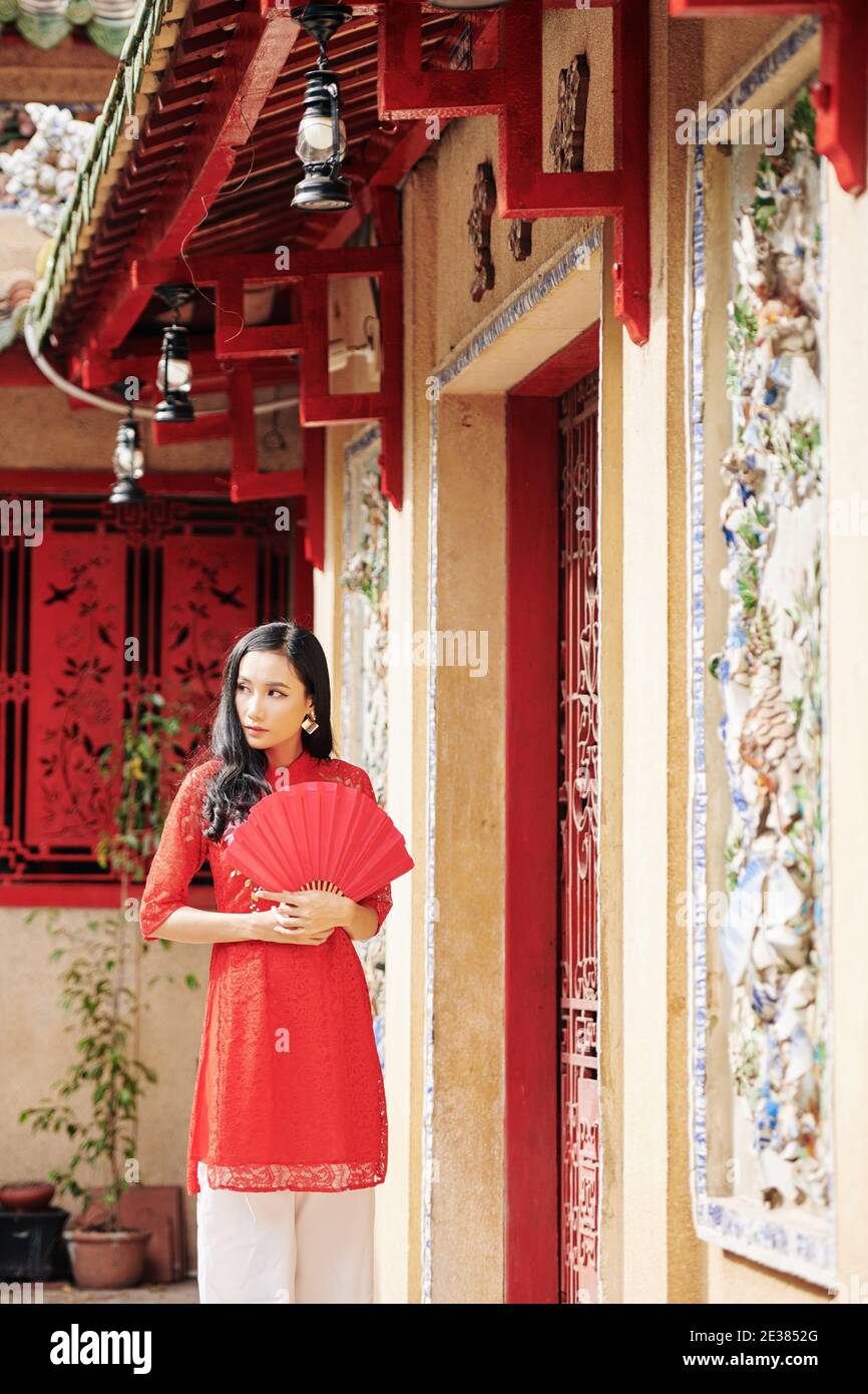 Bella giovane donna asiatica pensiva con ventola di carta rossa in piedi alle porte del tempio e guardando via Foto Stock