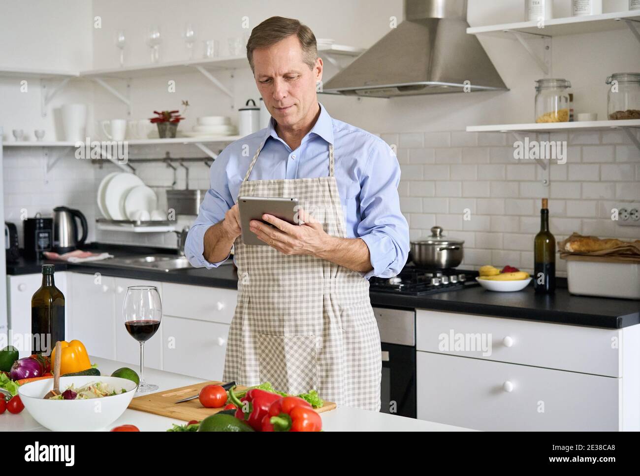 Uomo anziano anni 50 che indossa grembiule usando la tavoletta che prepara l'insalata in cucina. Foto Stock