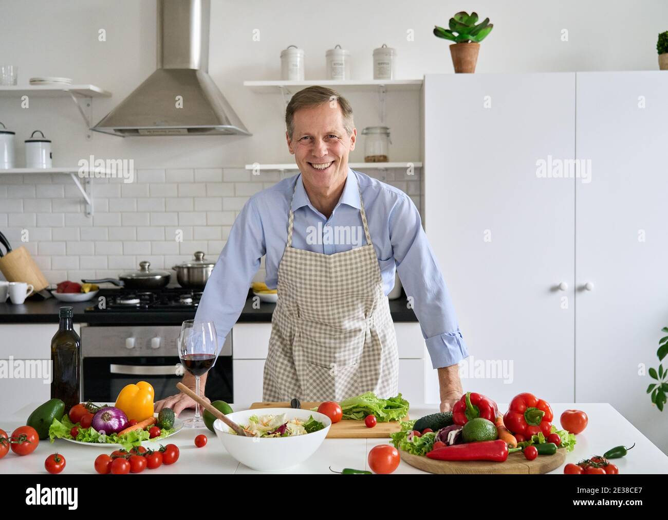 Sorridente uomo anni '50 che indossa grembiule in piedi in cucina tavolo preparare l'insalata. Foto Stock