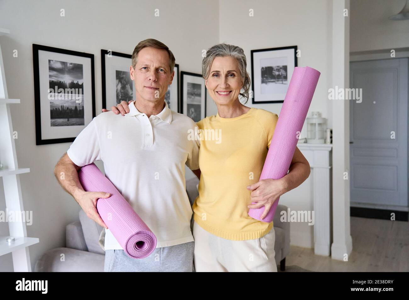 Felicemente calzata la vecchia coppia, guardando la macchina fotografica, tenendo i tappetini yoga a casa. Foto Stock