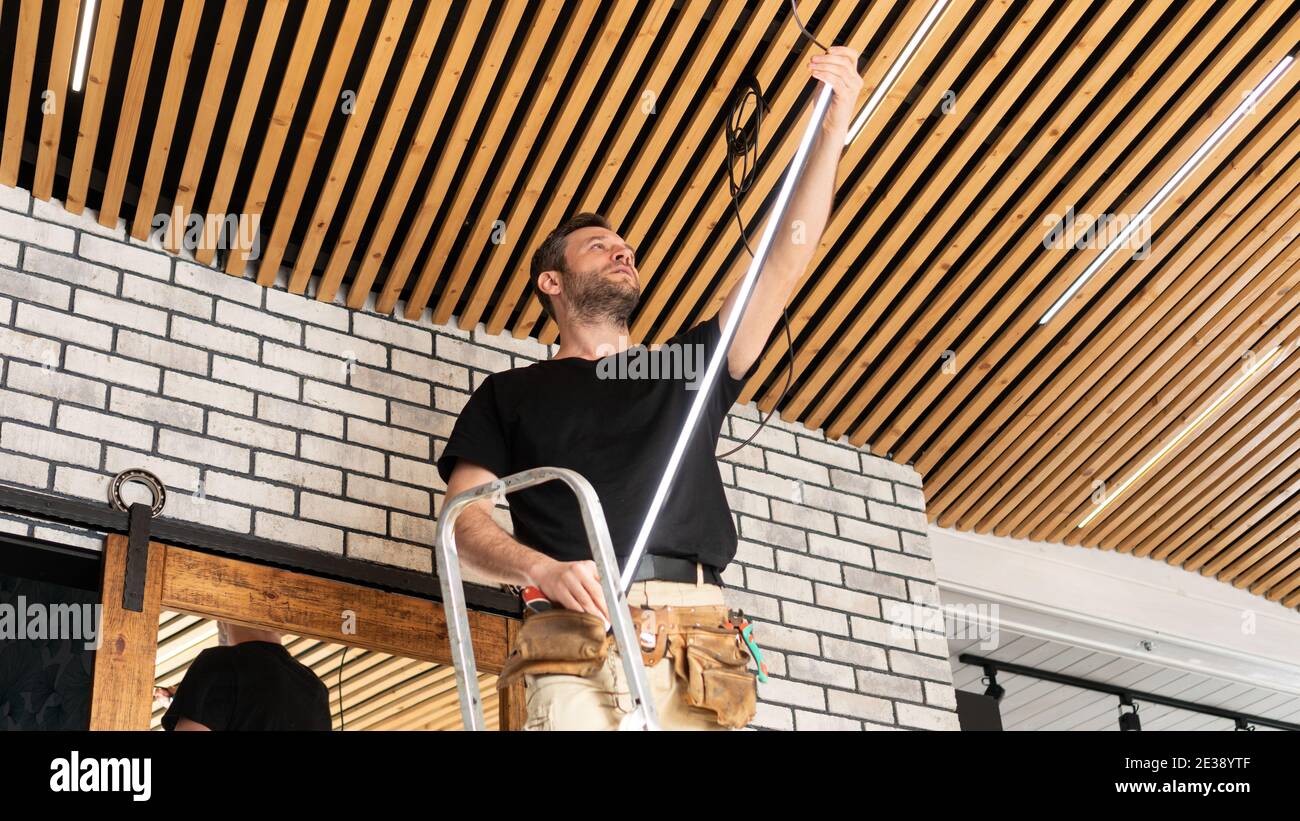Un elettricista maschio installa le lampade a striscia a LED in un soffitto con una tavola di pino. Un costruttore sta installando un soffitto in stile loft in un interno moderno. Assistenza per Foto Stock