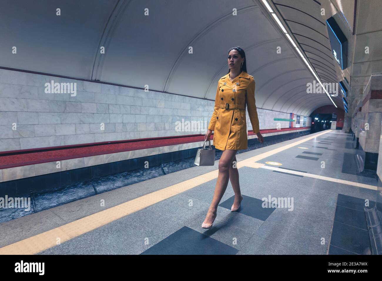 Camminando sulla piattaforma della metropolitana bella ragazza giovane con lunga bella gambe in un cappotto di primavera giallo e una borsa bianca o borsetta in mano Foto Stock