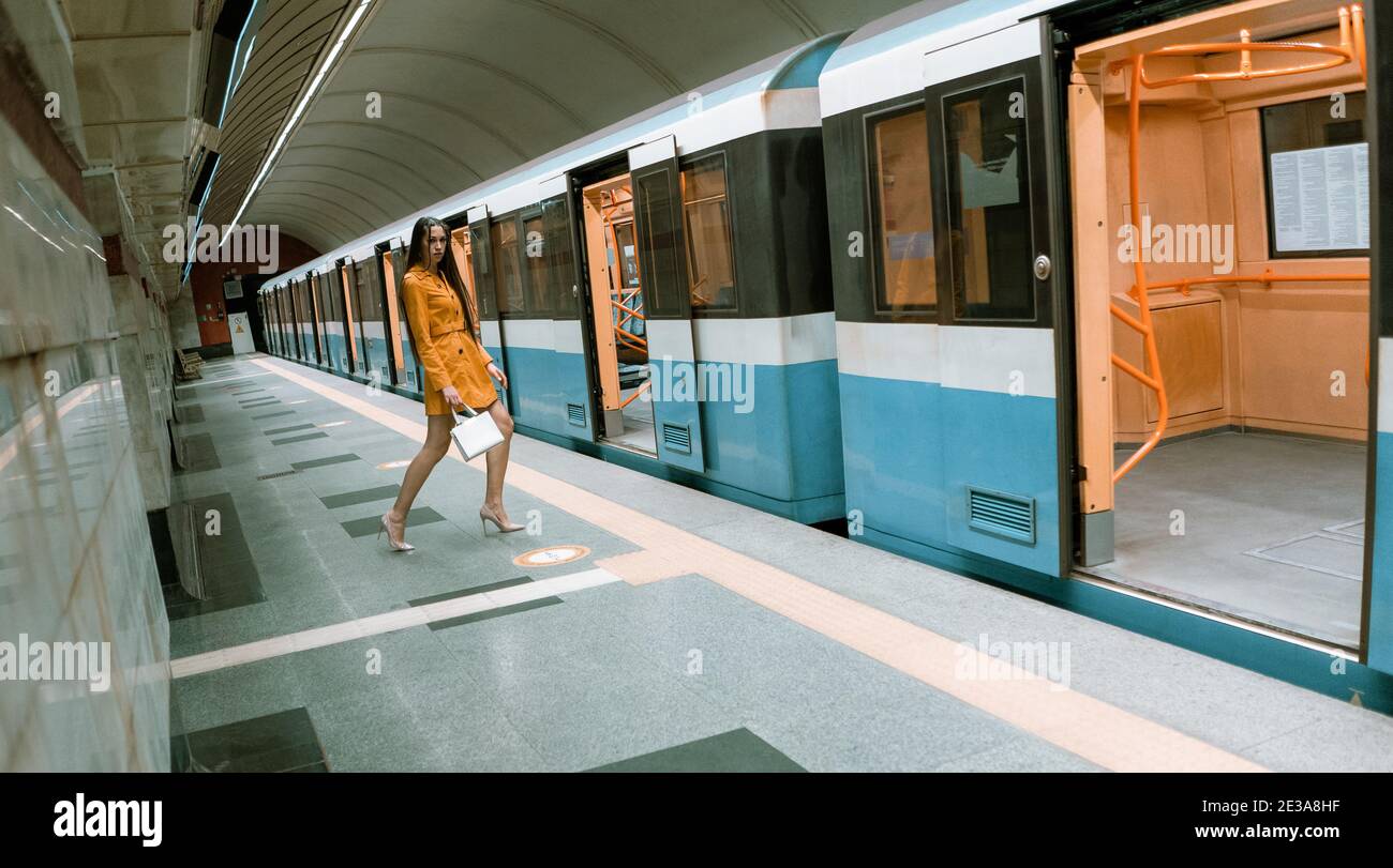 Posando di fronte ad una porta aperta di un treno della metropolitana vuoto, bella ragazza giovane con lunghe gambe belle in un cappotto di primavera giallo e una borsa bianca o. Foto Stock