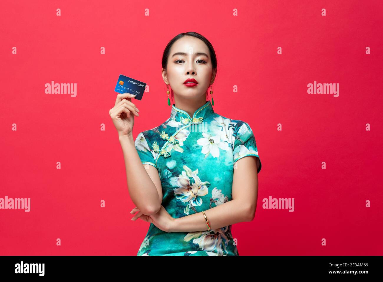 Bella donna asiatica che indossa abito orientale qipao mostrando carta di credito In mano su sfondo rosso studio per il nuovo anno cinese concetti di spesa Foto Stock