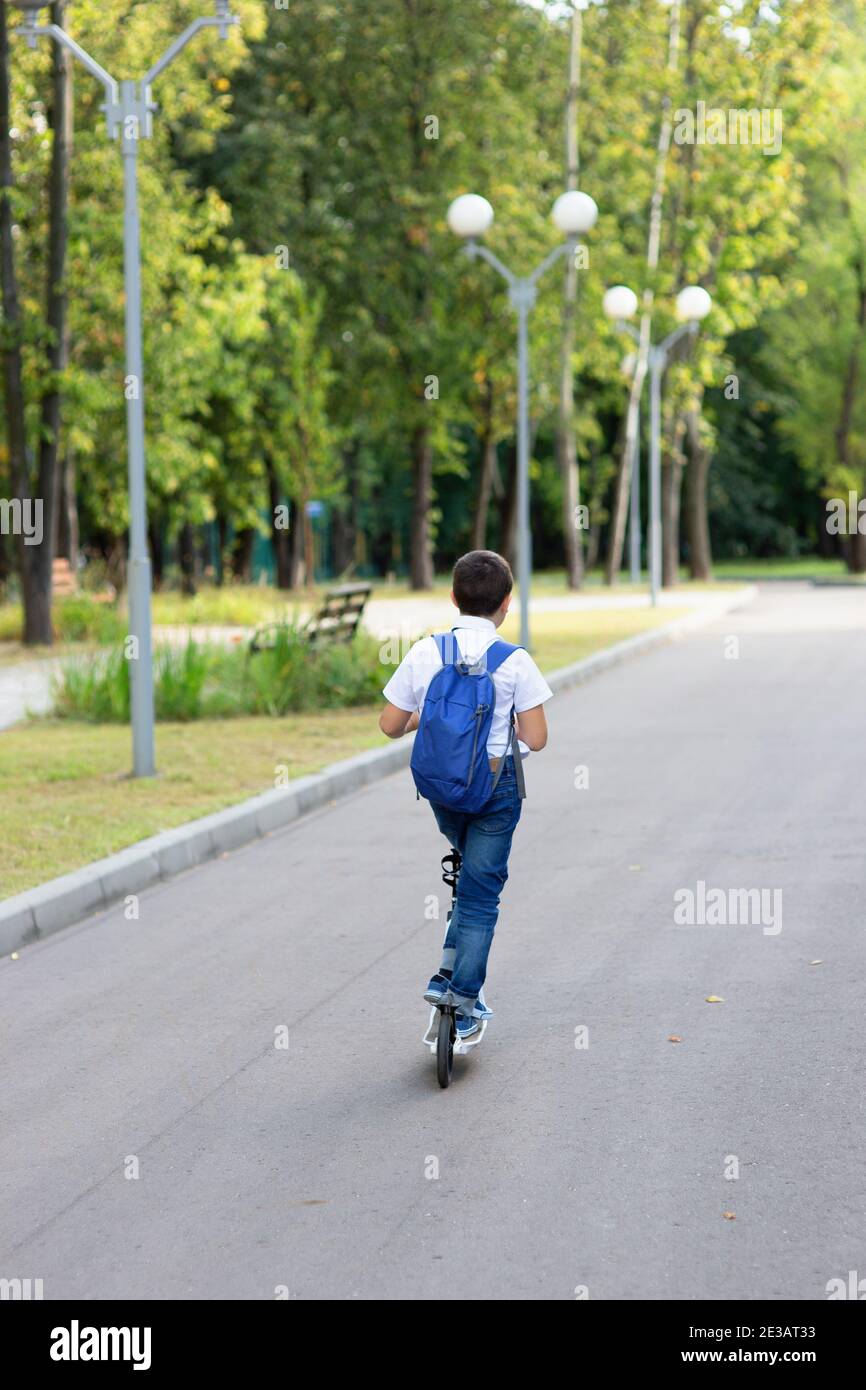 un bel ragazzo di stile e giovane in una camicia bianca, cravatta blu e uno zaino con uno scooter su una strada. vista posteriore Foto Stock