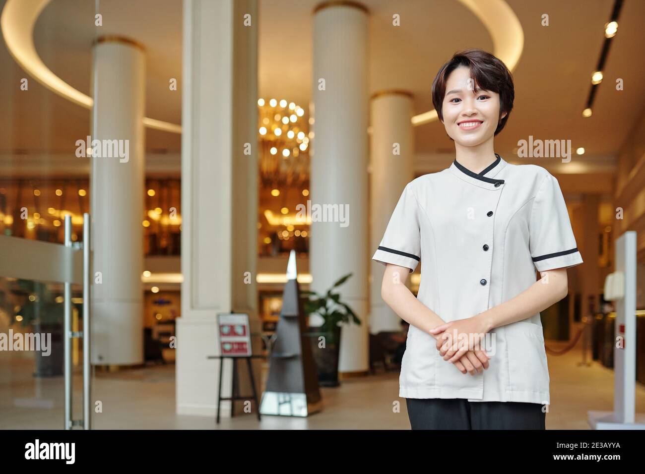 Un portiere vietnamita piuttosto giovane in uniforme che si trova all'ingresso dell'hotel e sorridendo alla macchina fotografica Foto Stock