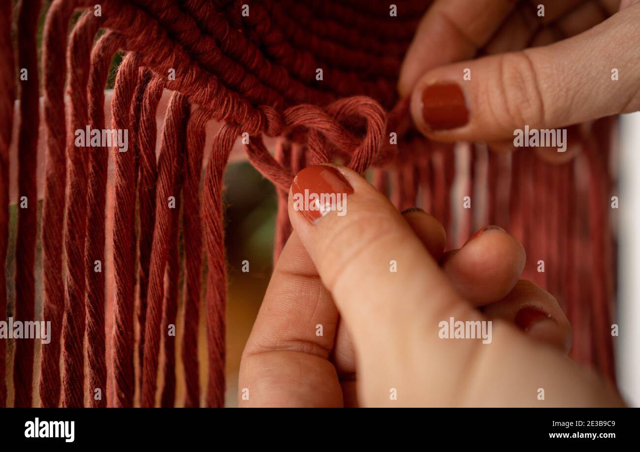 Mano femminile con smalto arancione per unghie fare nodi durante il lavoro un appendiabiti da parete in macrame Foto Stock