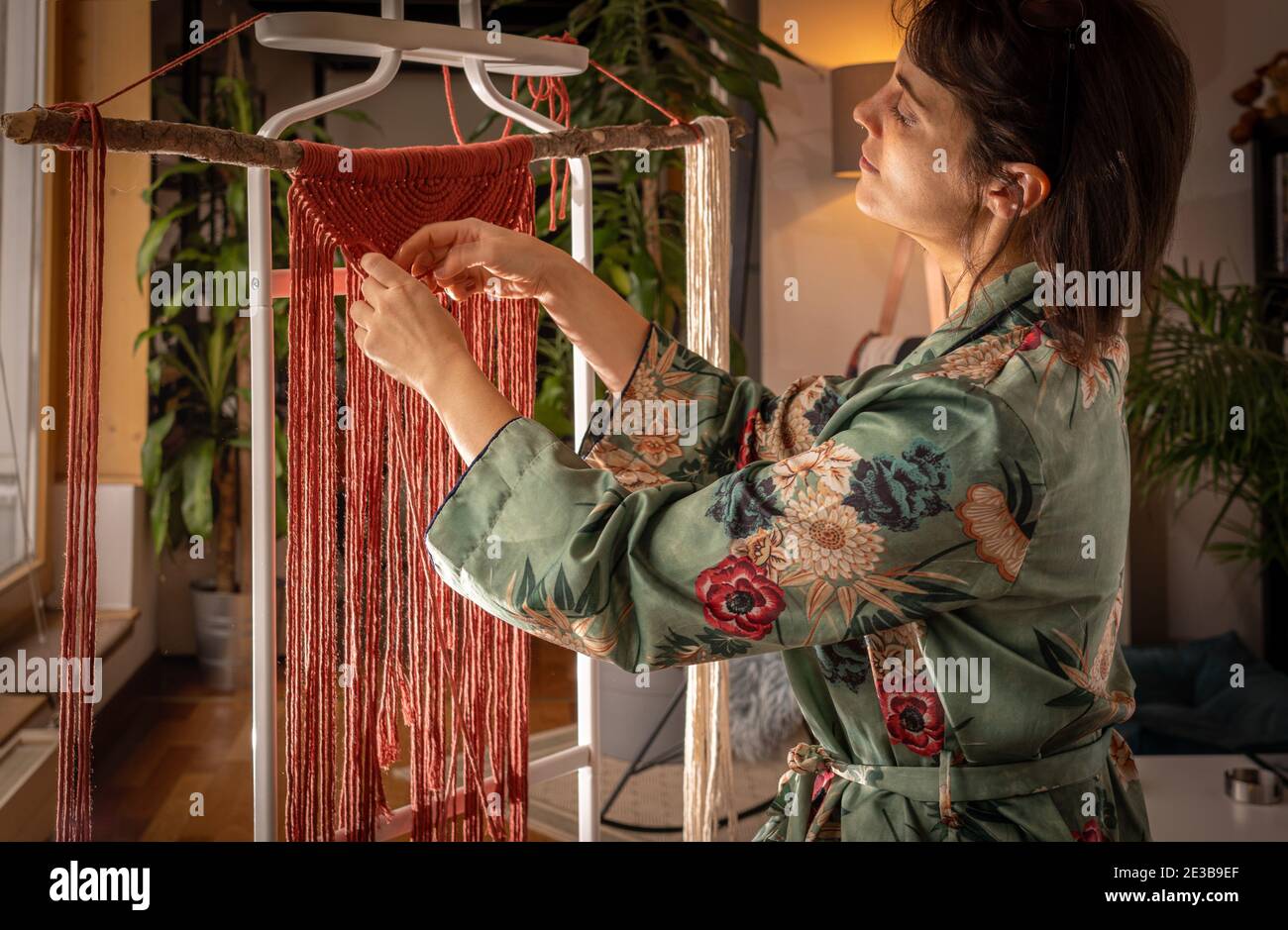 Una giovane donna che indossa un housecoat fa un'arte macrame lavoro durante una giornata di sole a casa Foto Stock