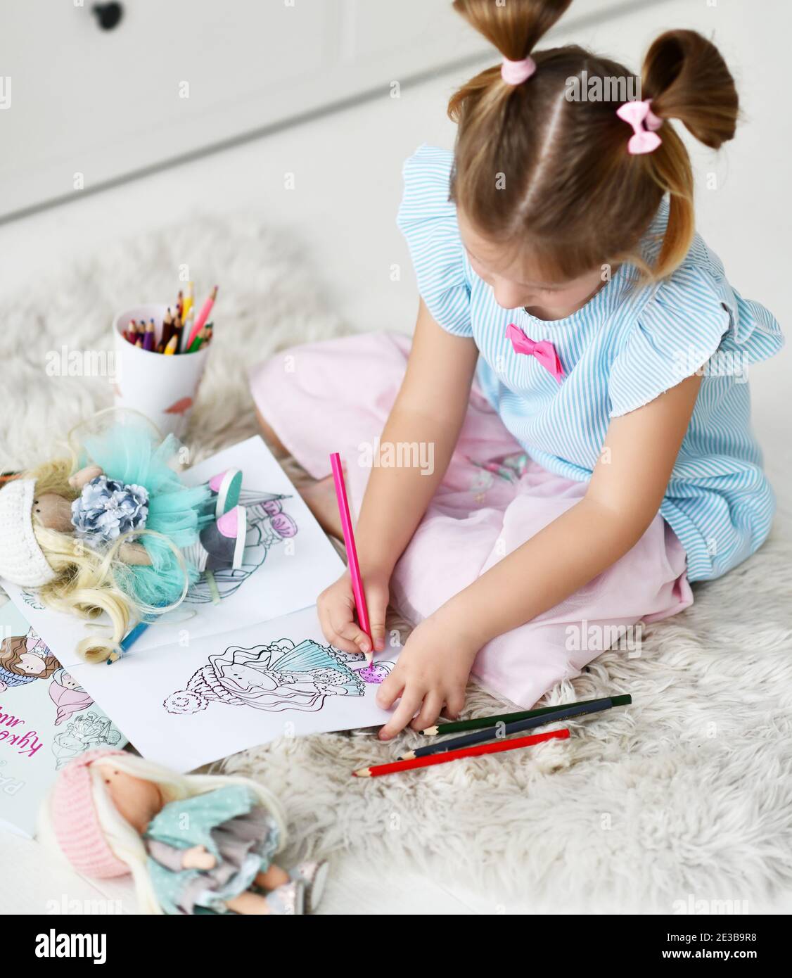 Ragazza di capretto con i panini divertenti si siede sul tappeto morbido a casa disegnando le immagini coloranti delle bambole con le matite colorate. Vista dall'alto Foto Stock