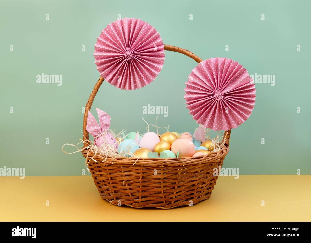 Cesto di vimini con colorate uova di Pasqua in colori pastello e decorato con fiori di carta. Buon biglietto d'auguri pasquale Foto Stock