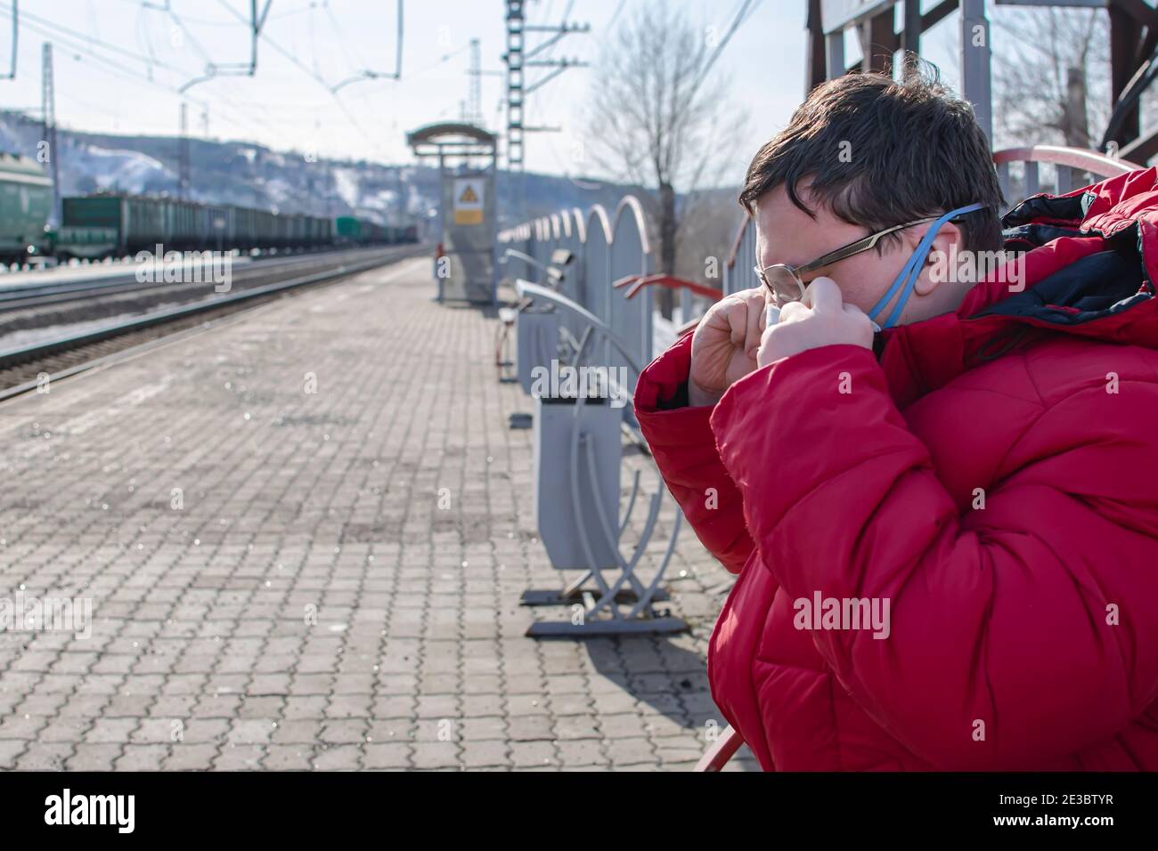 un uomo in attesa di un treno sulla piattaforma di una stazione ferroviaria regola una maschera medica anti-virus antibatterica su il suo volto Foto Stock