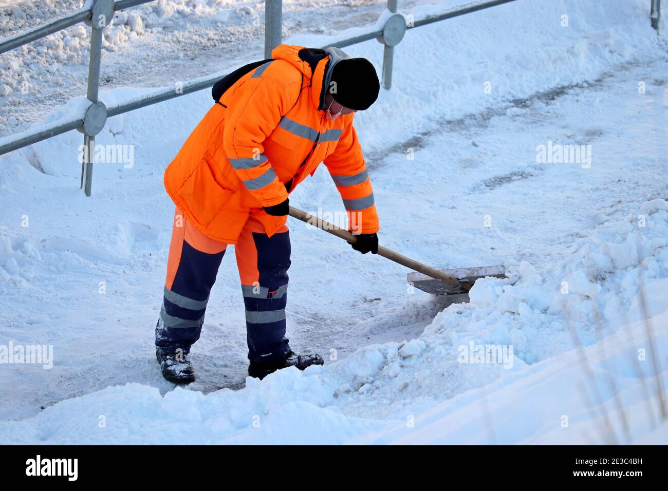 Rimozione della neve nella città invernale, pulizia della strada. Operaio dei servizi comunali in uniforme con una pala libera la neve su un marciapiede Foto Stock