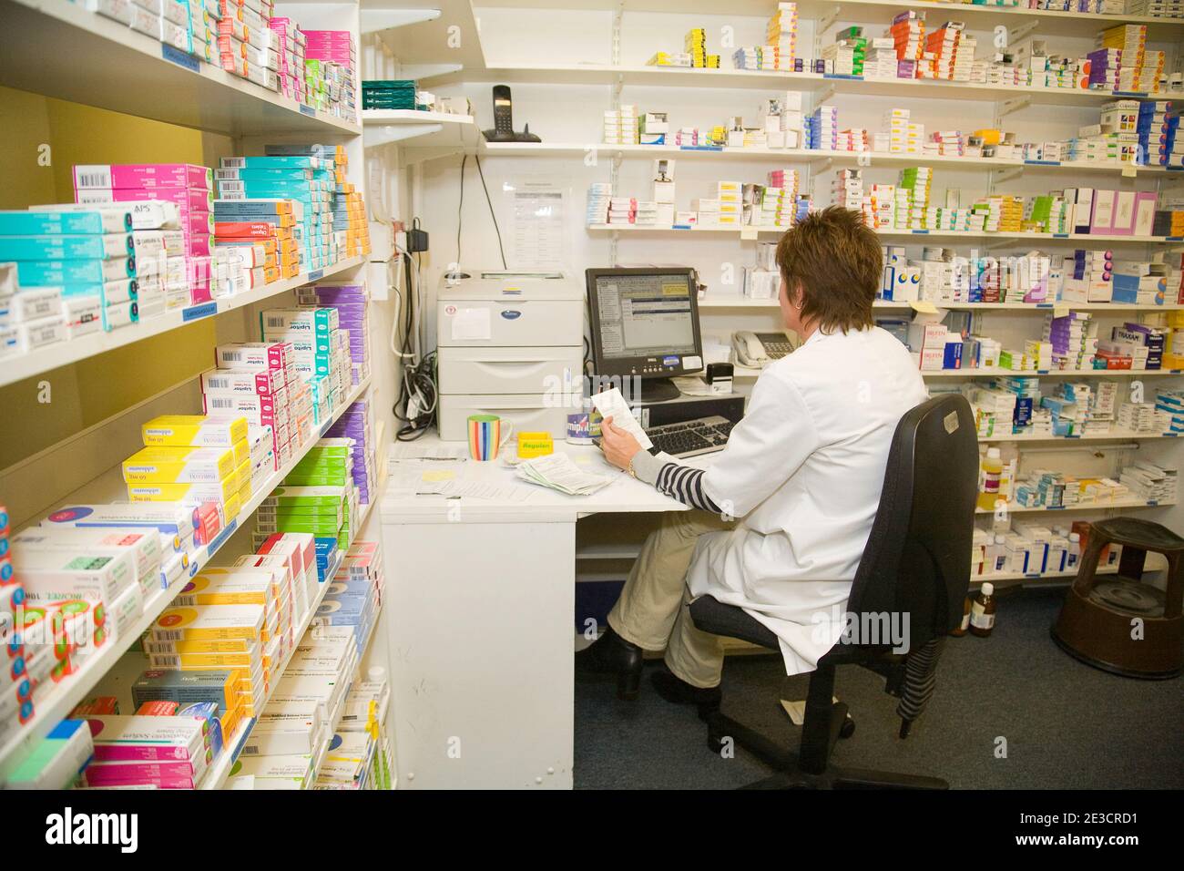 Pharmacy UK; Un farmacista seduto al computer della farmacia, circondato da scaffali di farmaci, Suffolk Inghilterra Regno Unito. Lavoratore britannico. Foto Stock