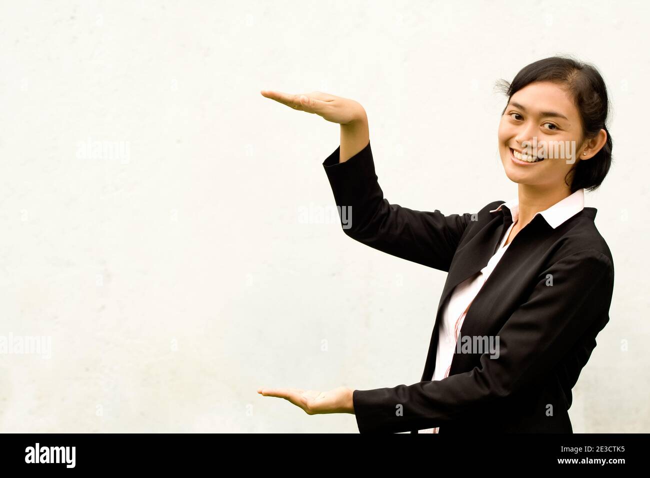 ritratto di giovane donna asiatica in tuta nera in piedi di fronte a uno sfondo bianco e alzando la mano mostrando un gap con le mani. Foto Stock