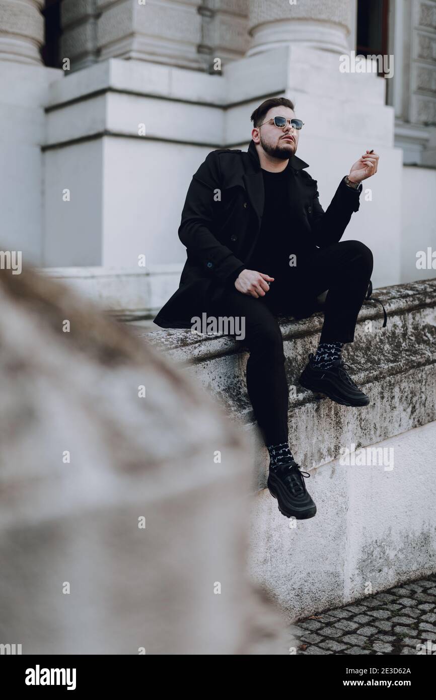 Lunghezza completa di un ragazzo elegante in occhiali da sole che riposano nel città mentre si fuma Foto Stock