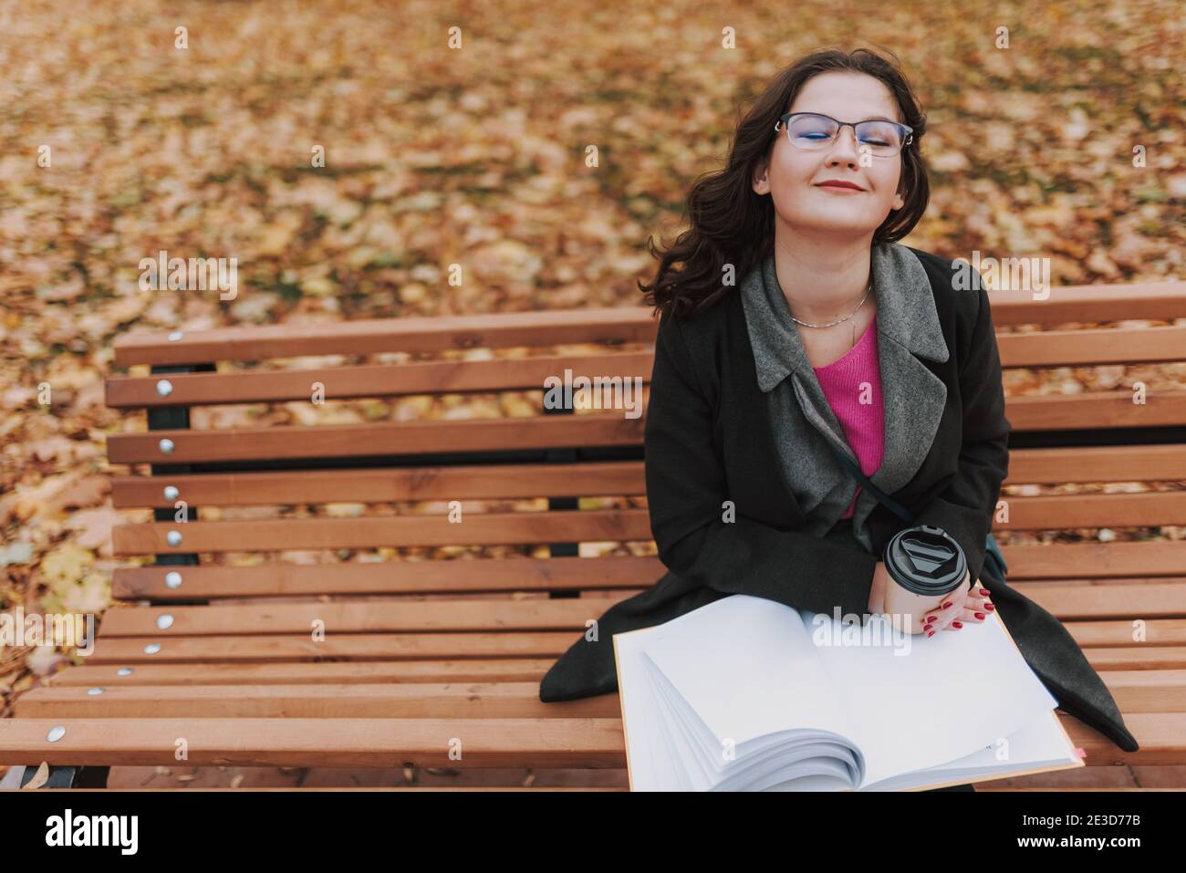 Vista dall'alto di una donna sorridente e attraente con gli occhi chiusi seduti sulla panchina e tenendo un libro, caffè nel parco Foto Stock